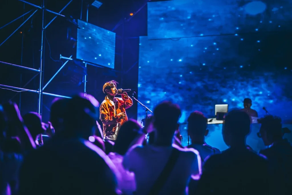 Live music performance featuring a singer on stage with large digital screens and a crowd watching in low light