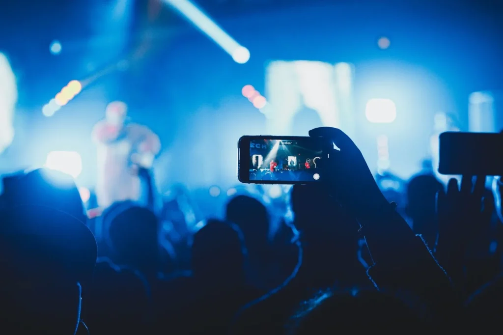 Audience member recording a live concert on a smartphone with stage lights and performers visible on screen