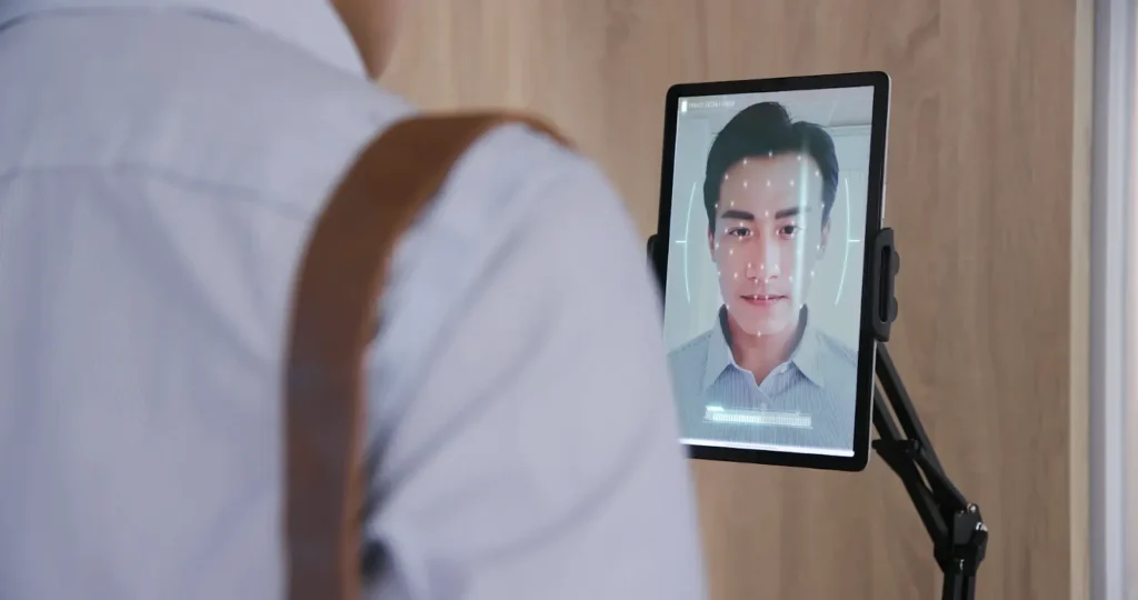 Person standing in front of a tablet using facial recognition technology, with a digital face scan overlay visible on the screen.