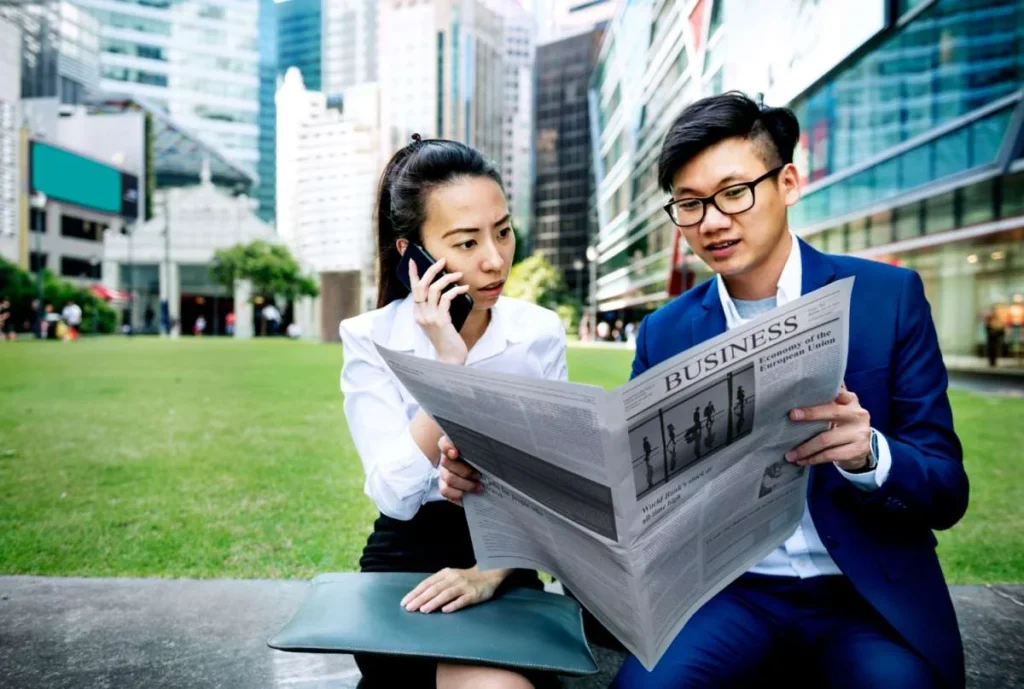 Two professionals sitting outdoors reading a business newspaper and discussing current affairs